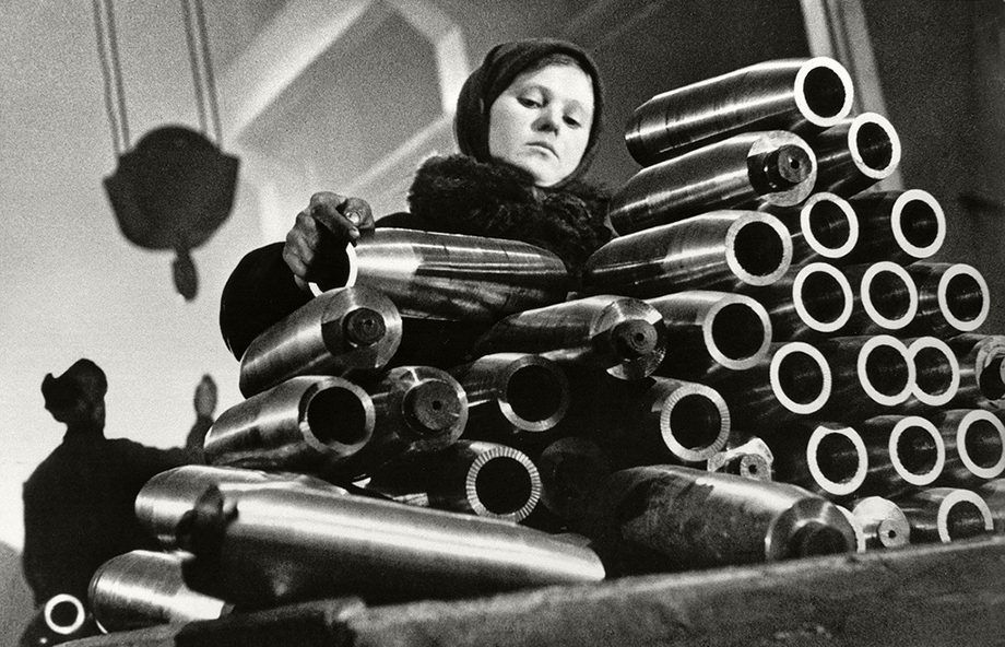Девочка в заводском цехе укладывает болванки снарядов.